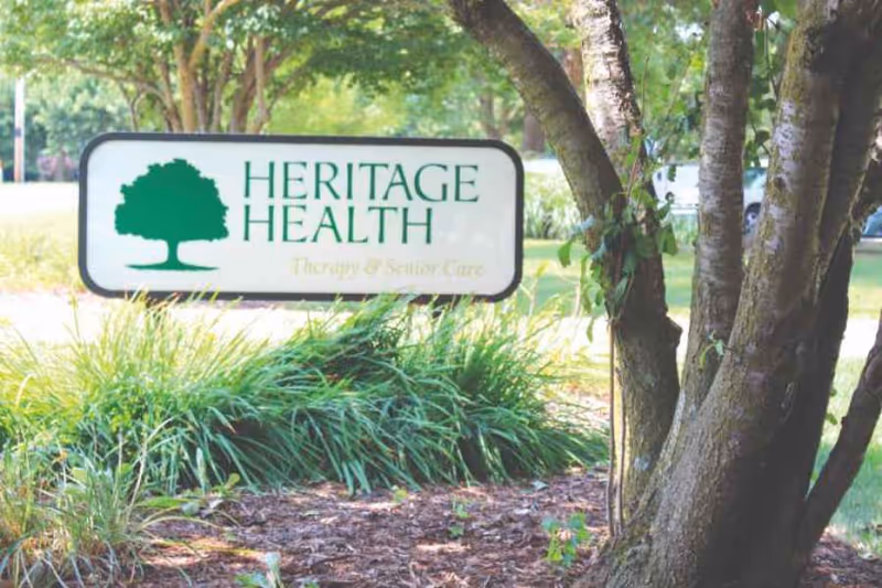 Outdoor view of a green and white sign for Heritage Health Therapy & Senior Care, surrounded by grass, plants, and trees in a landscaped area.
