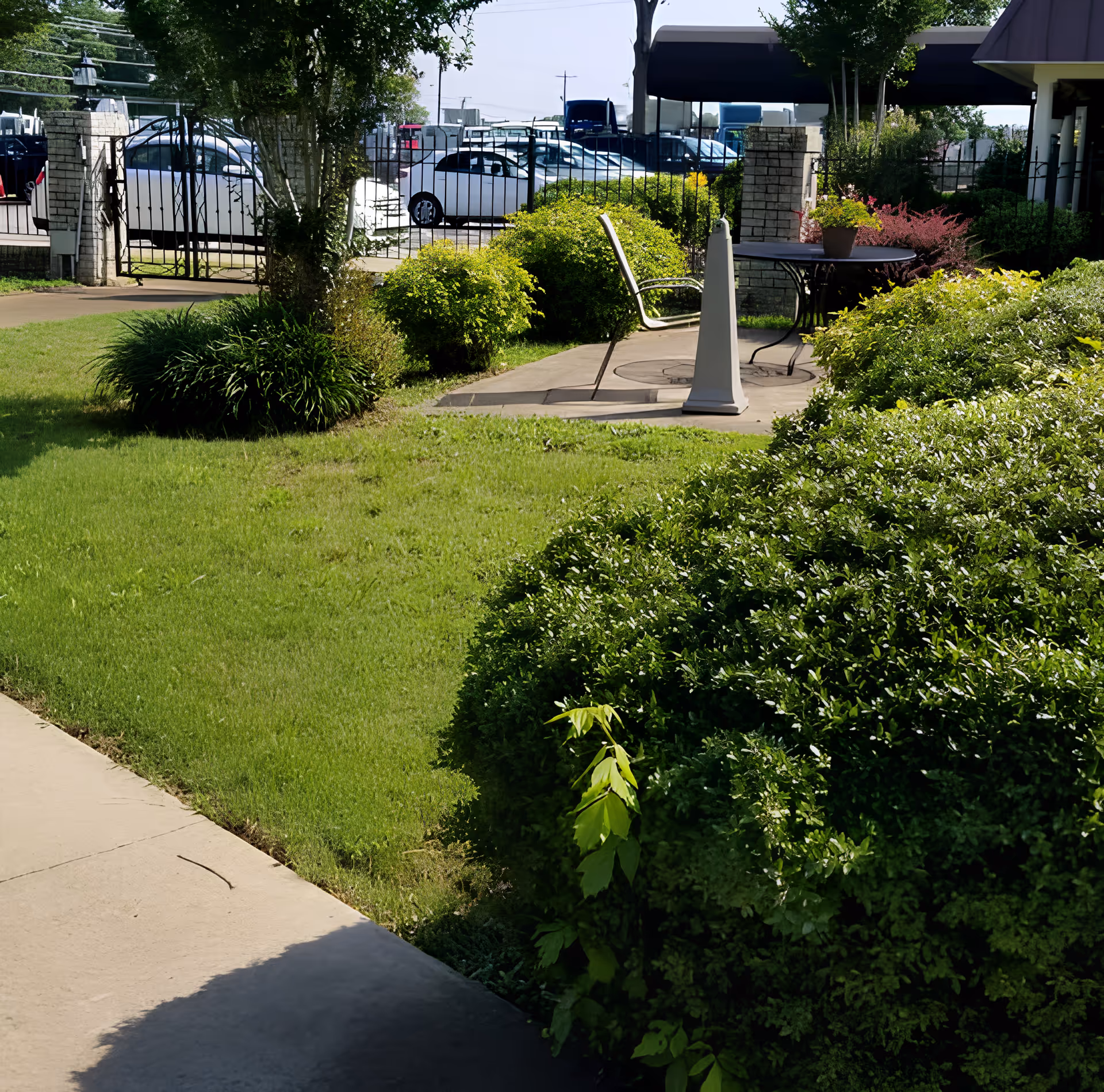 Small outdoor patio with a table and chairs surrounded by green lawn and shrubs in front of a gated parking area.