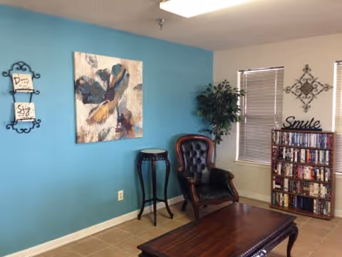 A cozy living room area with a blue accent wall featuring a large floral painting and two small framed quotes. There is a dark wooden coffee table in the foreground, a tall plant in the corner, a vintage-style armchair with tufted black leather upholstery, a small round side table, and a bookshelf filled with DVDs. The walls are light beige, and there are two windows with blinds partially closed. A decorative wall piece and the word 'Smile' are displayed above the bookshelf.