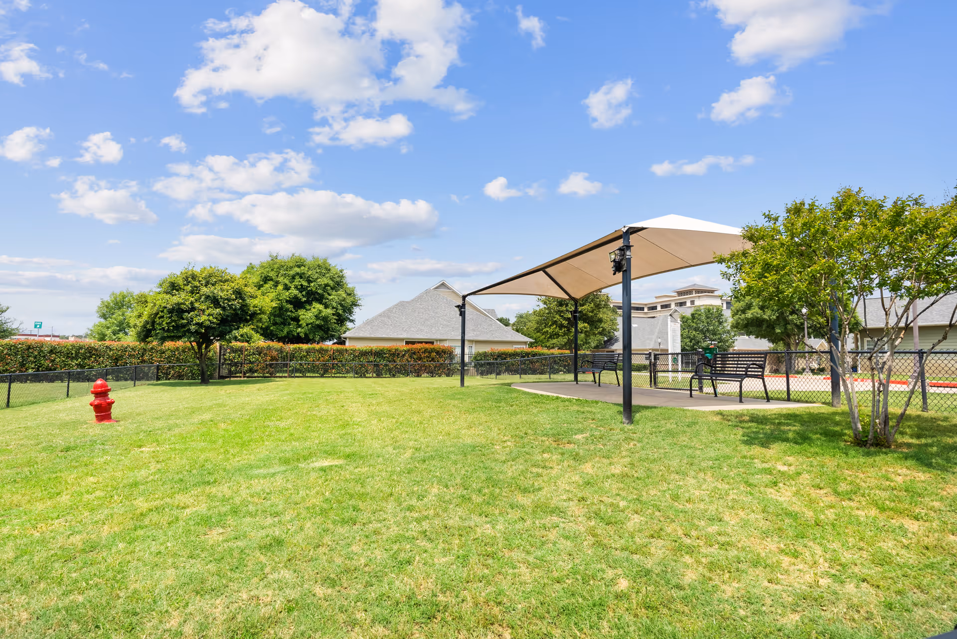 Grassy outdoor area with a shaded pavilion and benches, trees, and a red fire hydrant near a fence.