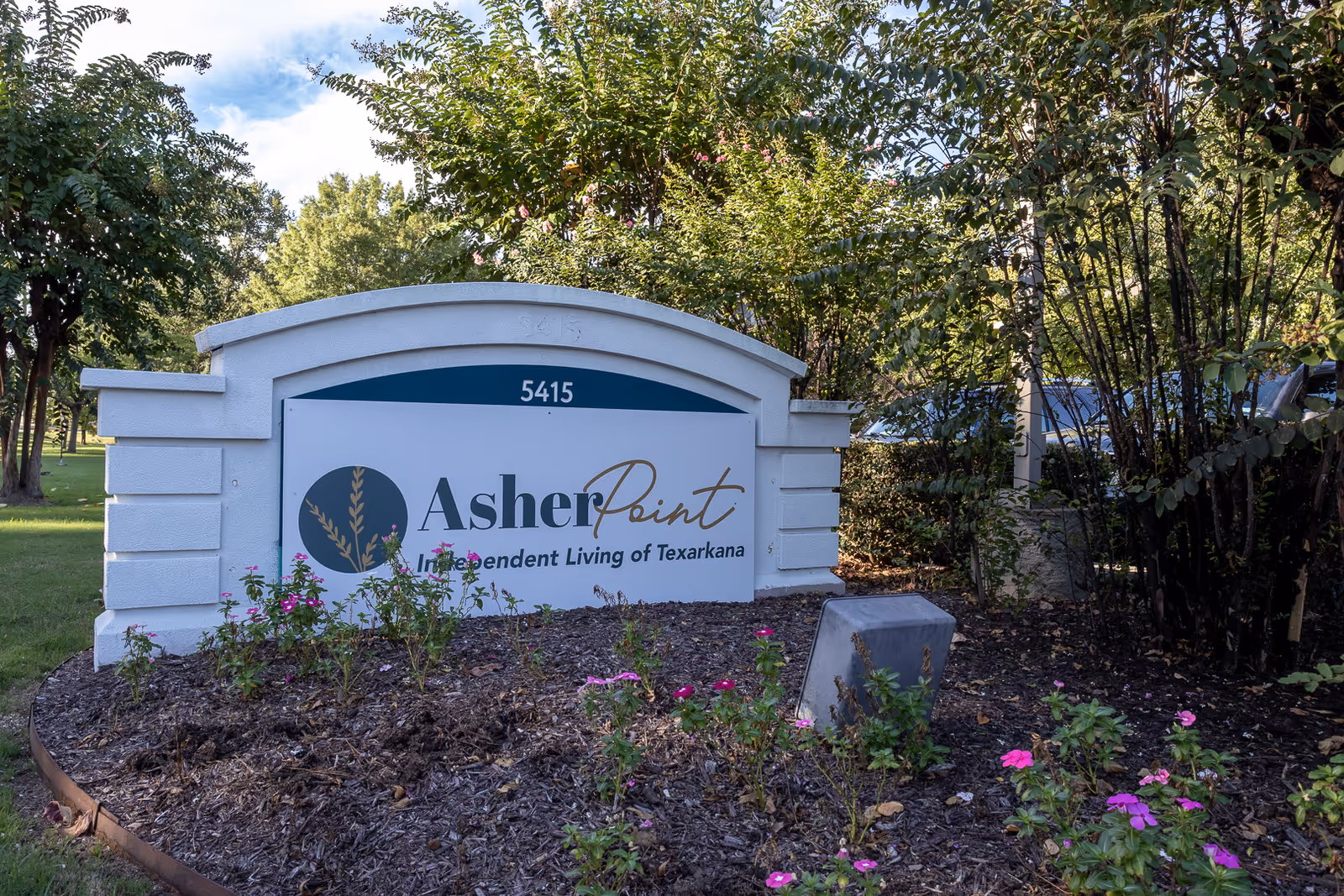 Outdoor view of a white stone sign for Asher Point Independent Living of Texarkana surrounded by green trees and pink flowers in a landscaped garden area.