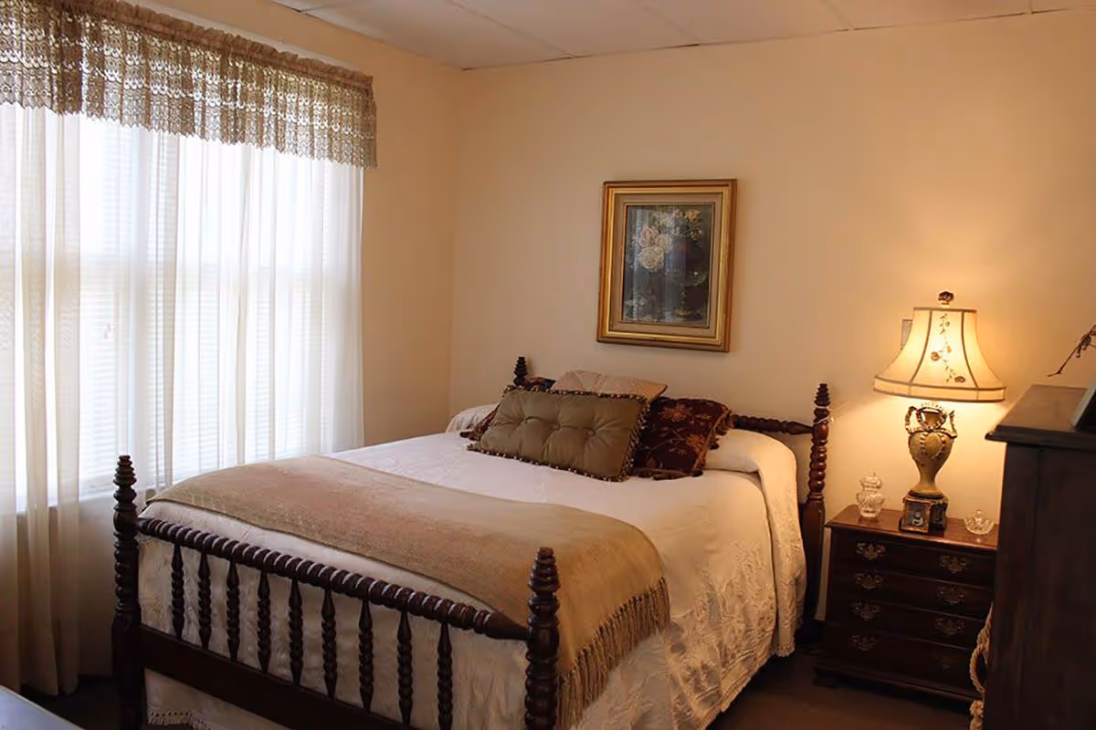 A cozy bedroom with a wooden bed frame, white bedding, and decorative pillows. A beige throw blanket is draped across the foot of the bed. Next to the bed is a wooden nightstand with a decorative lamp and small glass items. A framed floral painting hangs on the wall above the bed. Light filters softly through sheer curtains on a large window.