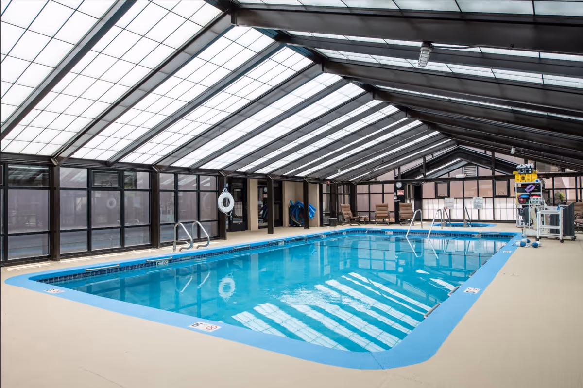 Indoor swimming pool with clear blue water under a glass roof structure. The pool area has beige flooring, metal handrails, life rings on the walls, and pool equipment on the right side. There are several lounge chairs against the far wall.