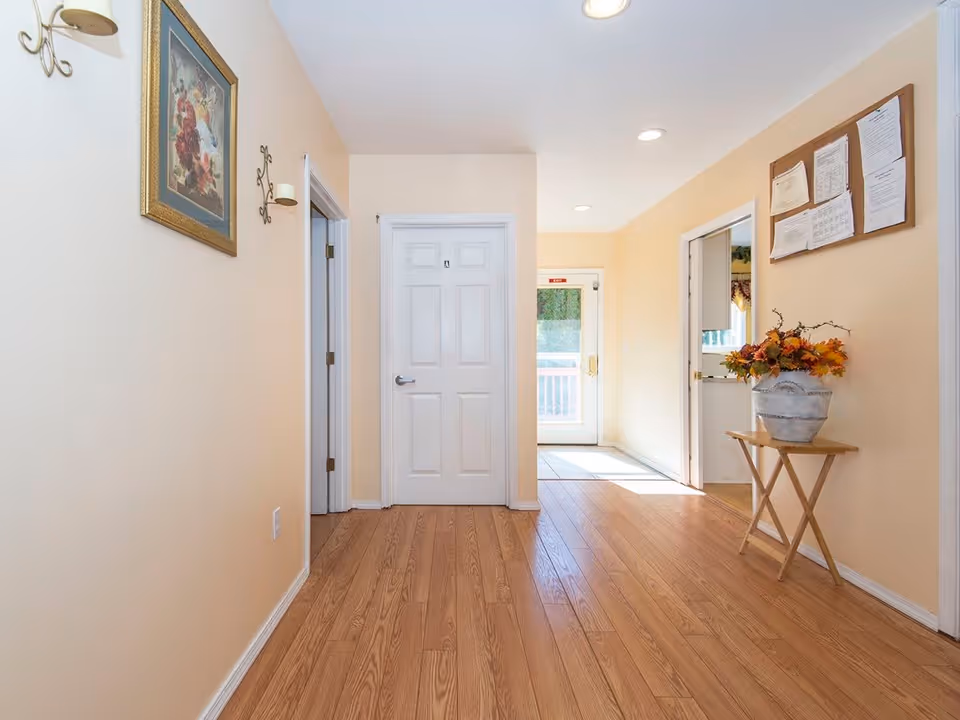 A bright hallway with light wood flooring and cream-colored walls. There are three white doors, one in the center with a restroom sign, and two open doorways on either side. A small wooden table with a vase of autumn leaves is on the right side, below a bulletin board with papers pinned to it. A framed floral painting and two wall-mounted candle holders are on the left wall. Natural light comes through a glass door at the end of the hallway.