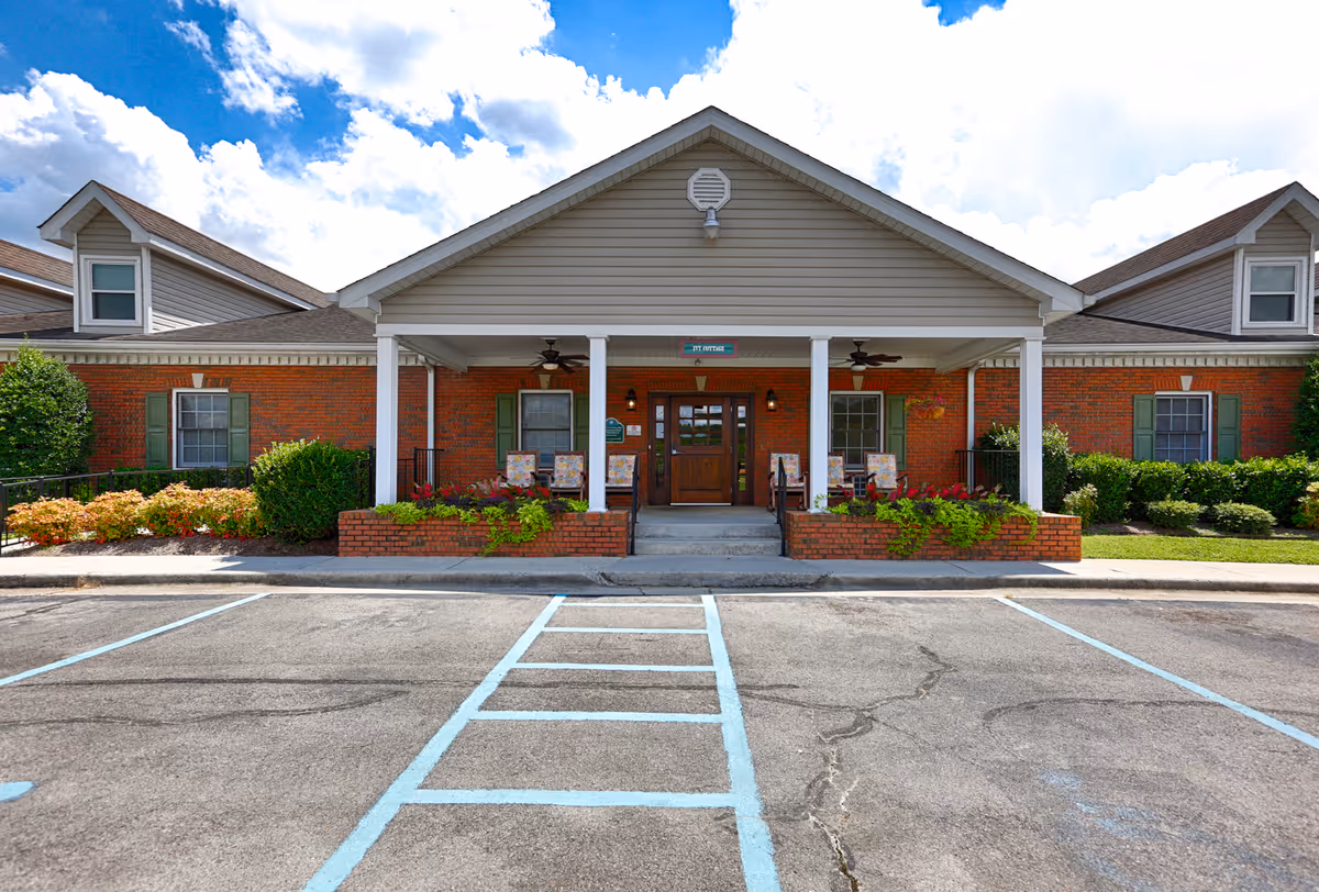 Front exterior view of a single-story brick building with a covered entrance supported by white columns. The entrance has a wooden door with windows on either side, surrounded by green shutters and landscaping with bushes and flowers. There are chairs on the porch and a parking area with marked spaces in front.