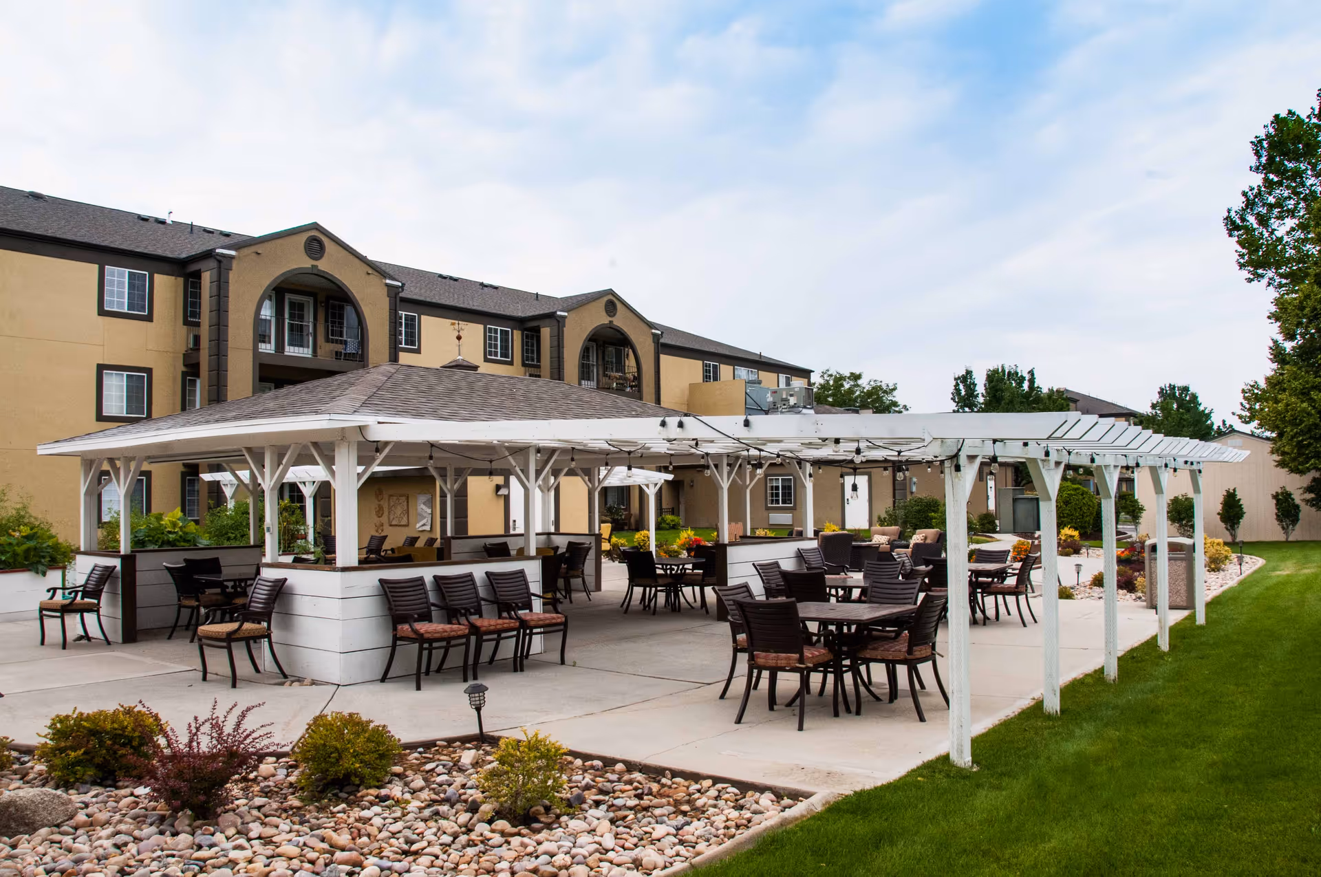 Outdoor patio area at Cedarwood at Sandy with multiple tables and chairs under a white pergola and covered seating area, surrounded by landscaping with rocks, bushes, and green grass, with a multi-story building in the background under a cloudy sky.