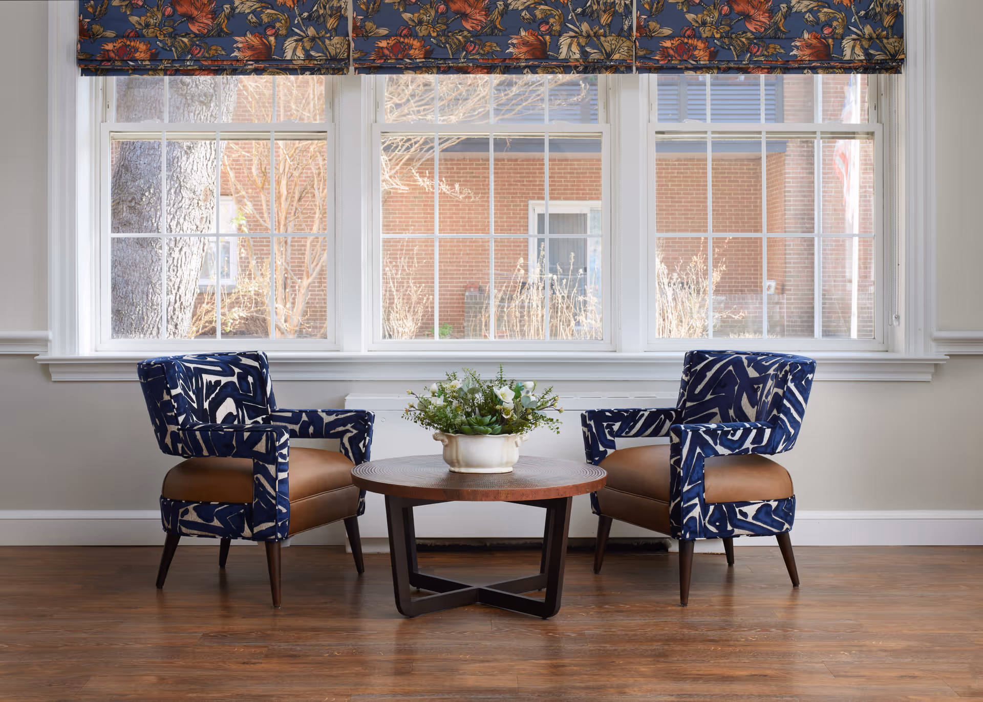 Two patterned armchairs facing a round wooden coffee table with a potted plant in front of large multi-pane windows.