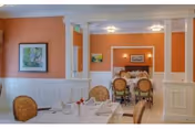 Bright dining room with orange walls, white wainscoting and columns, set tables and upholstered chairs leading into a secondary dining area.