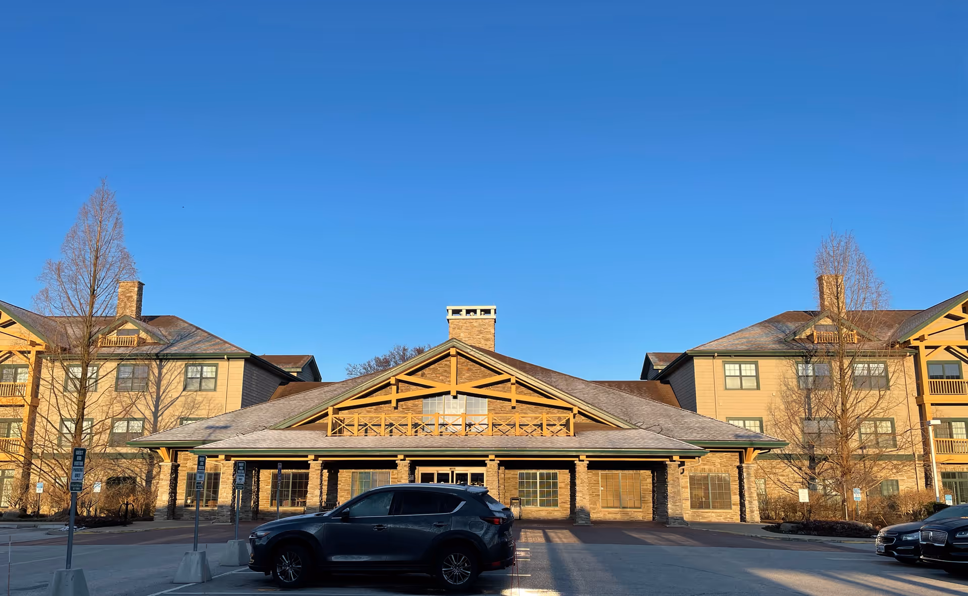 Front exterior view of Deer Creek Lodge, a large building with a peaked roof, stone pillars, and wooden architectural details. There are two three-story wings on either side with balconies and windows. Several cars are parked in the parking lot in front of the building under a clear blue sky.