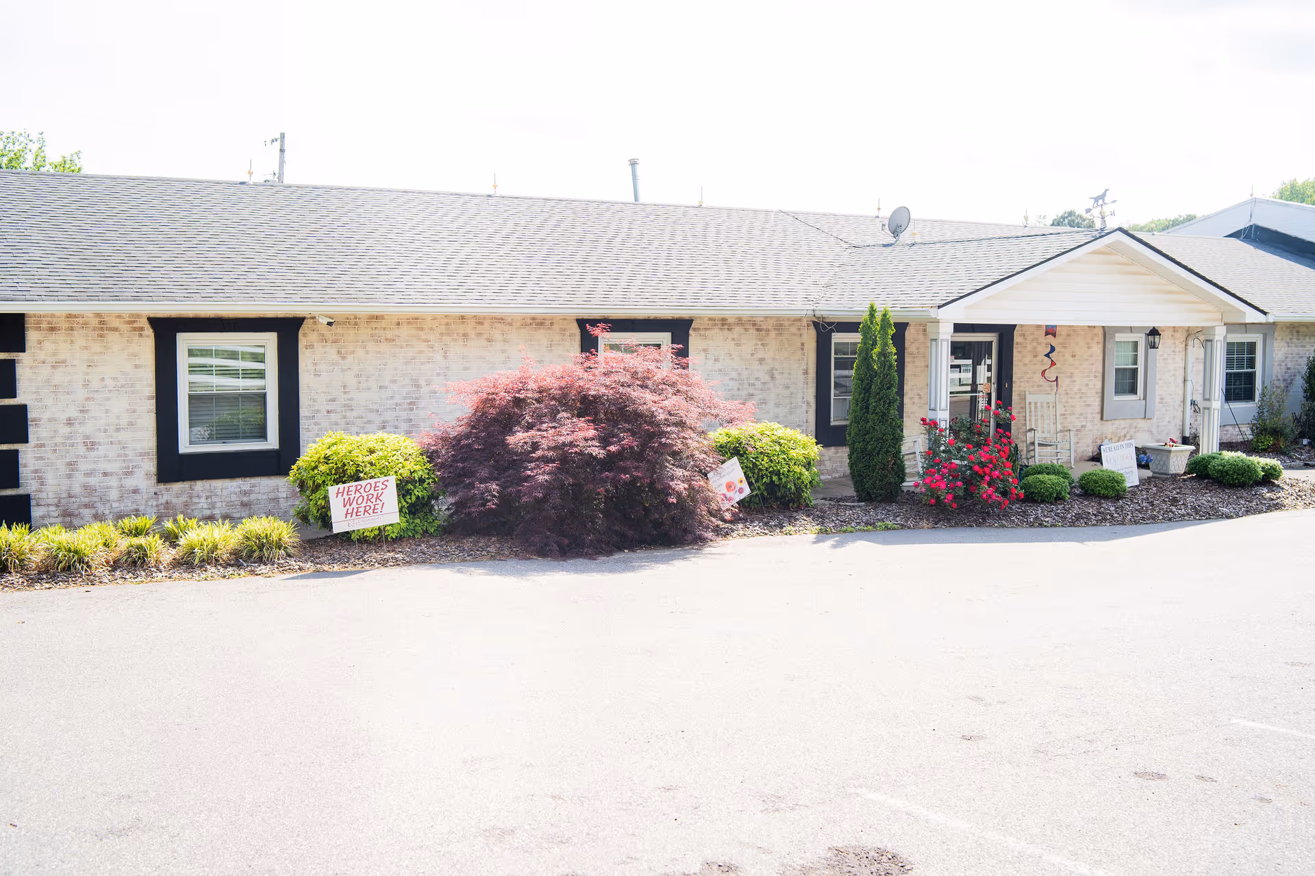 Exterior view of a single-story brick building with black window frames and a gray shingled roof. There are various shrubs and flowering plants in front of the building, including a large reddish bush and red flowers near the entrance. Two signs are visible in the landscaping, one reading 'HEROES WORK HERE!' and another partially visible. The entrance has a small covered porch with white columns and two rocking chairs.