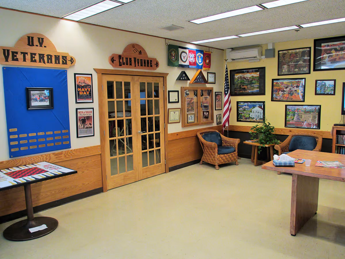 A bright communal room with wicker chairs, framed pictures, an American flag, wall displays, and double wooden doors.