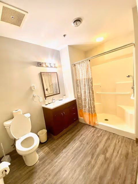 A clean and well-lit bathroom featuring a white toilet with the lid open, a dark wood vanity with a white countertop and a mirror above it, and a shower area with a patterned shower curtain. The floor is covered with wood-like vinyl flooring and the walls are painted light beige.