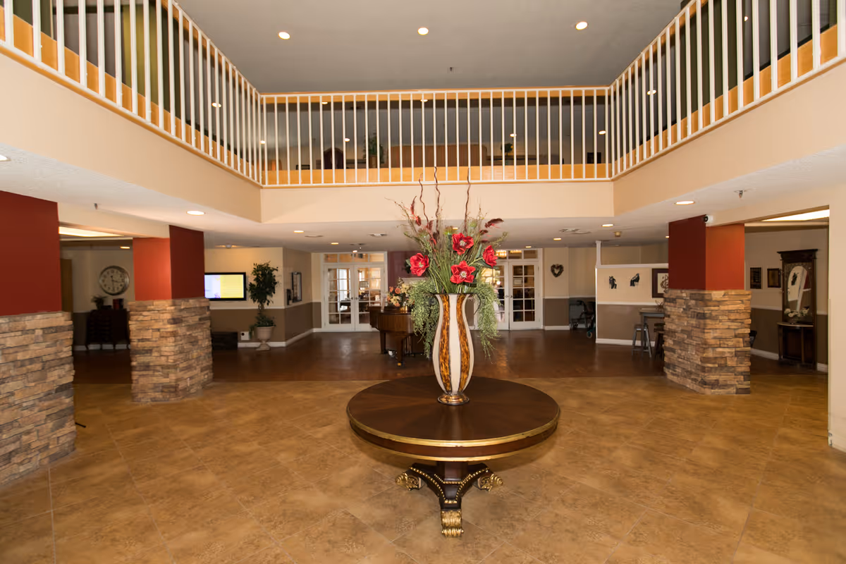 Spacious senior living facility lobby with a round wooden table in the center holding a large decorative vase filled with red flowers and greenery. The lobby features tiled flooring, stone pillars with red accents, and a second-floor balcony with white railings overlooking the area. There are double glass doors at the back and various pieces of furniture and plants around the space.