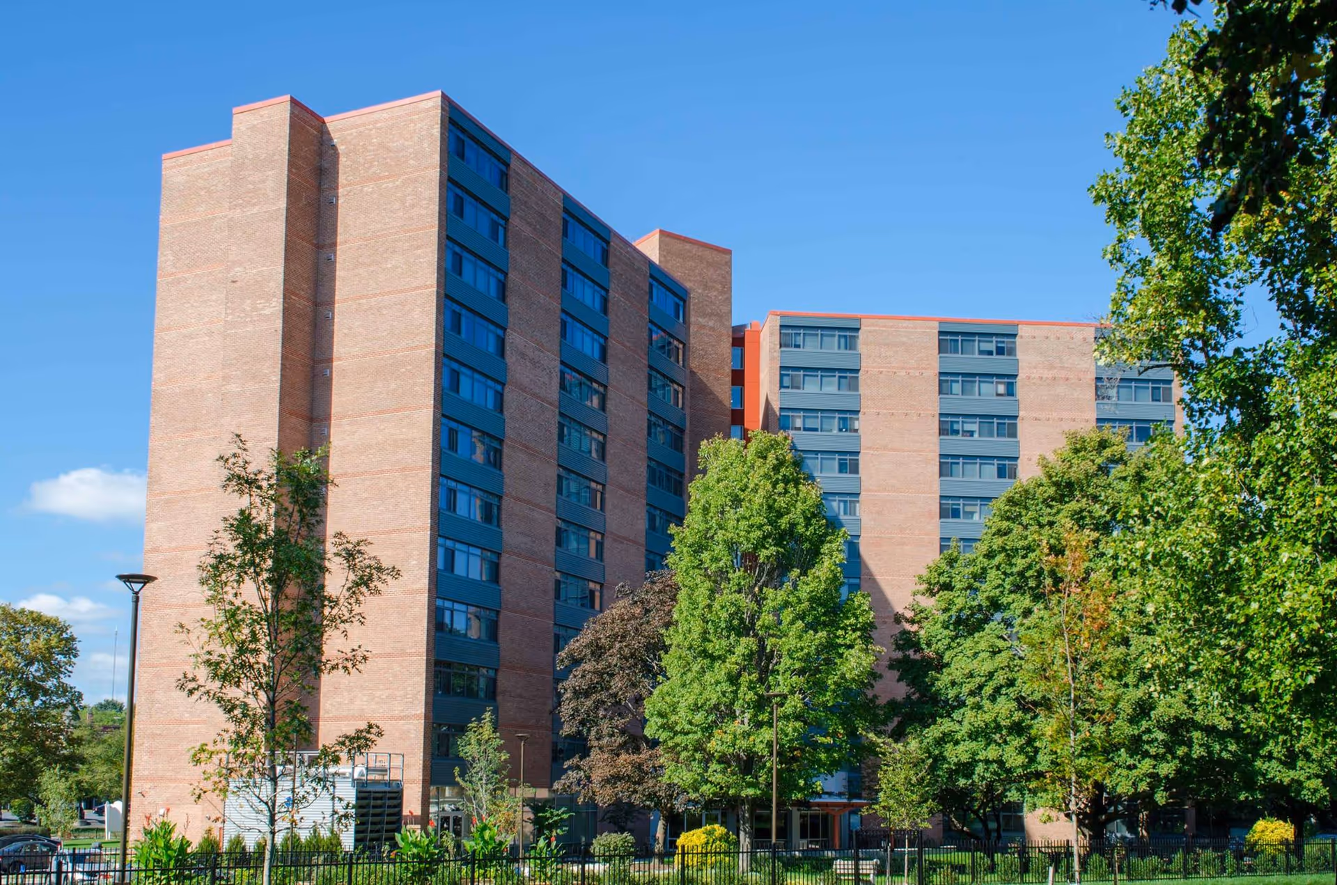 Exterior view of a multi-story brick apartment building with large windows, surrounded by green trees and a clear blue sky.