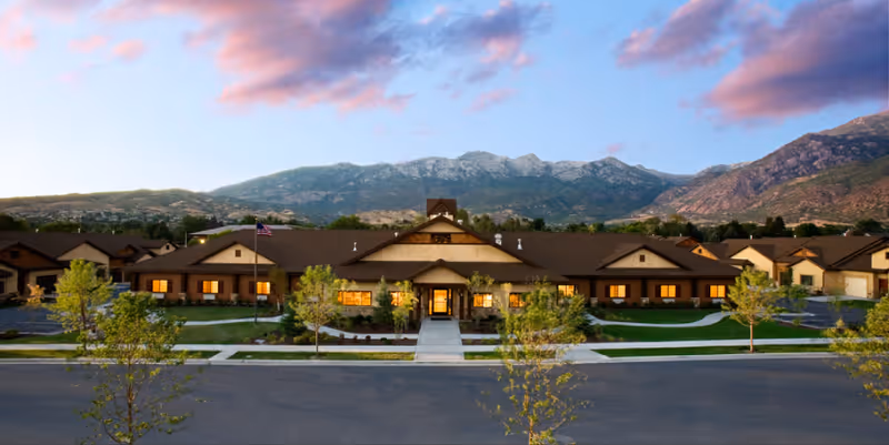 Exterior front view of River Meadows Senior Living facility at dusk with warm lights glowing from windows, a flagpole with an American flag, landscaped greenery, and mountains in the background under a partly cloudy sky.