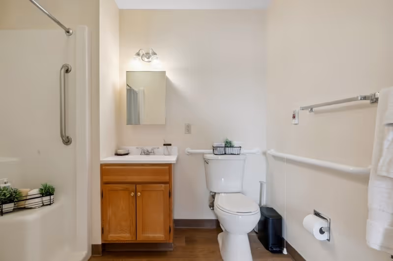 A clean senior living bathroom with a wooden vanity cabinet, a sink with a mirror and light fixture above it, a white toilet with a small basket on top, grab bars on the walls for support, a towel rack with a white towel, and a shower area with a grab bar and toiletries.