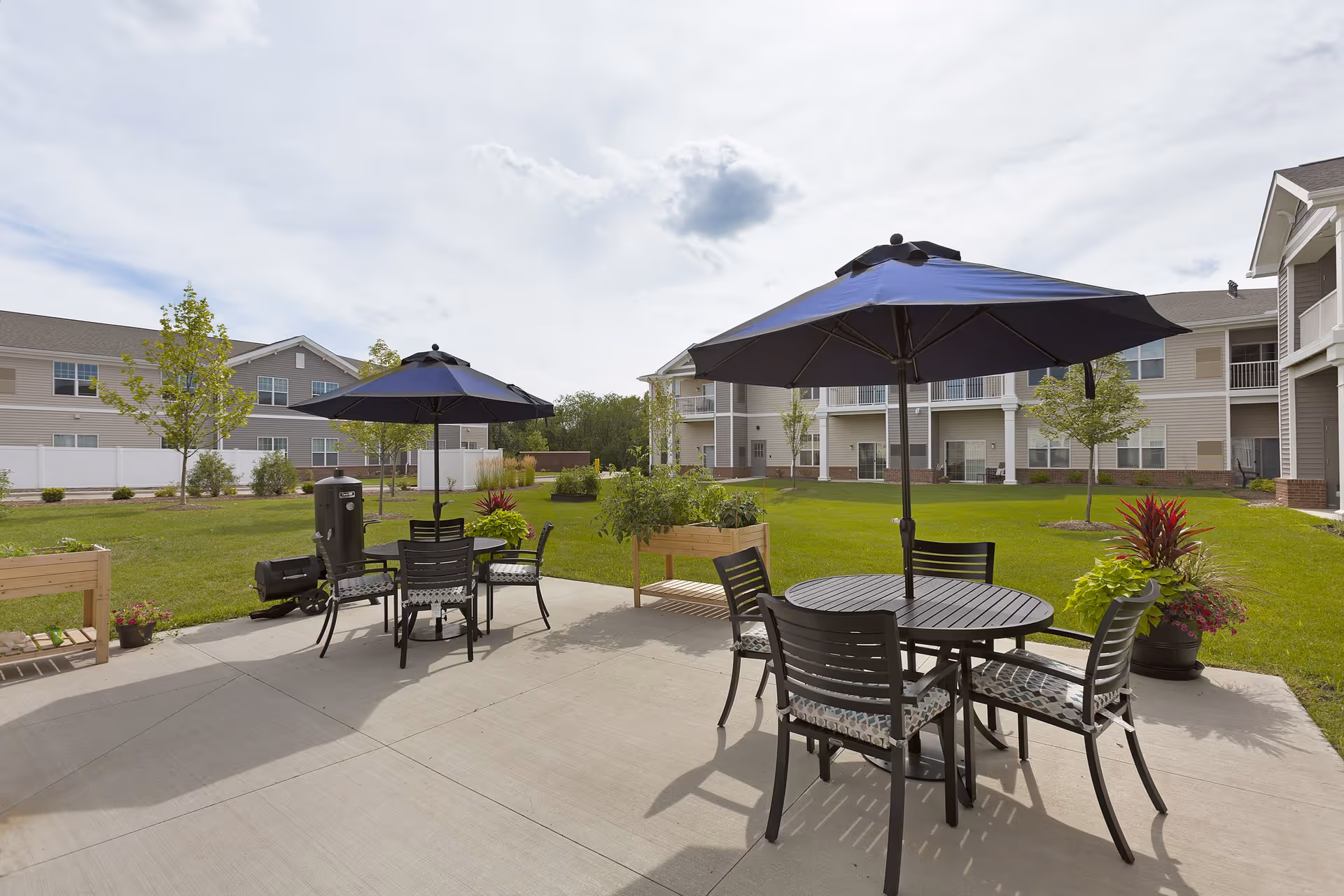 Outdoor courtyard with round patio tables, chairs and blue umbrellas on a concrete patio overlooking a grassy lawn and multi-story senior living buildings.