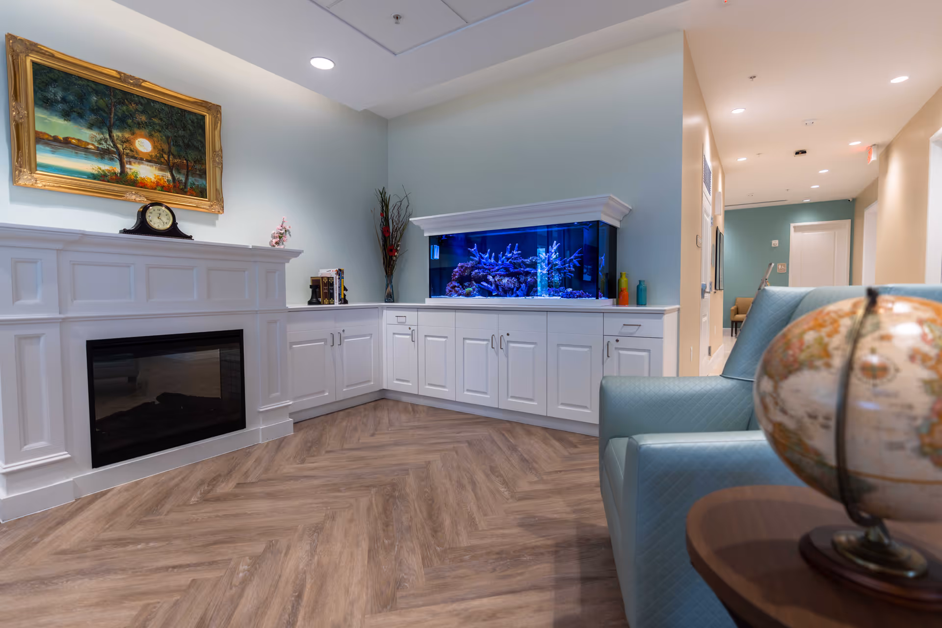 A cozy interior corner of a senior living facility featuring a white fireplace mantel with a clock and a framed painting above it, a large built-in aquarium with blue lighting, white cabinetry, a light blue armchair, and a globe on a wooden side table. The floor has a wood-patterned design and the walls are painted in soft pastel colors.