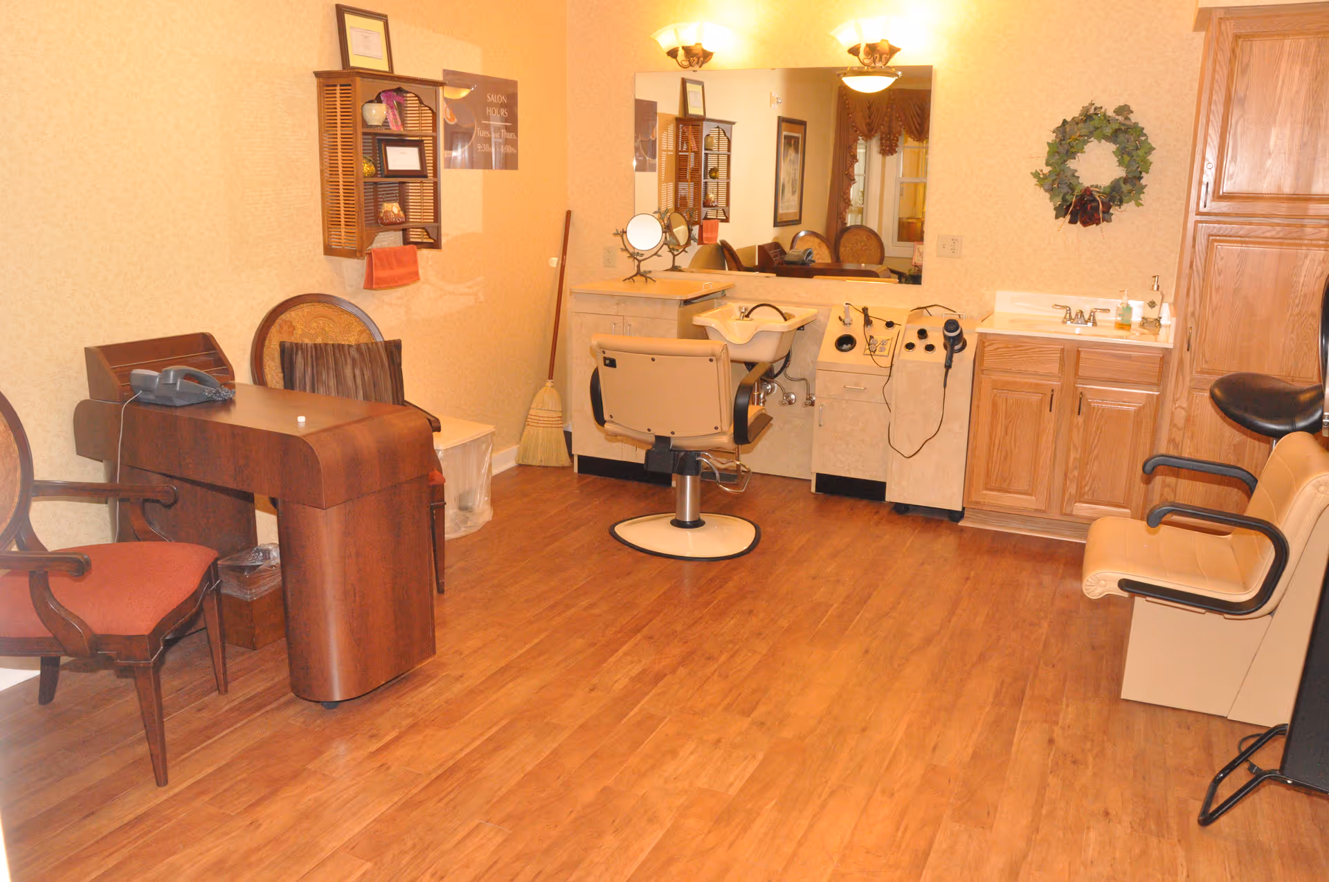 Interior of a salon room with a styling chair in front of a sink and mirror, wooden cabinets, a small desk with a telephone, two chairs, a broom in the corner, and a decorative wreath on the wall.