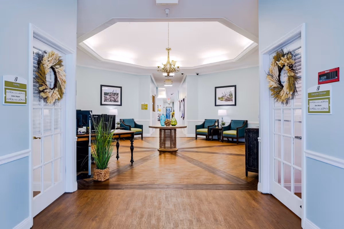 Bright reception lobby with seating, a central table under a chandelier, and double glass doors decorated with wreaths.