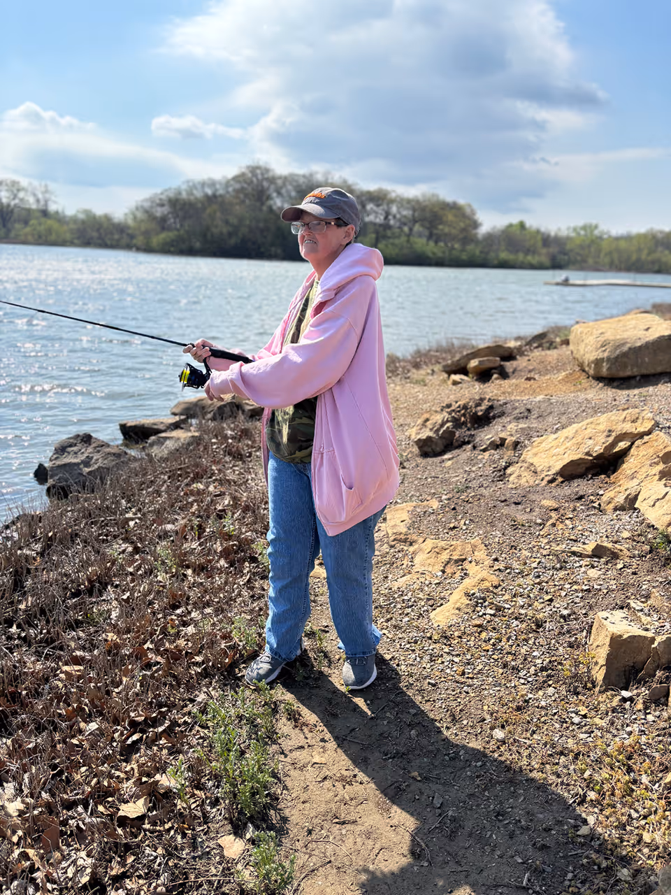 A person wearing a pink hoodie, blue jeans, and a cap is fishing by the edge of a lake on a sunny day. The shoreline is rocky and there are trees in the background under a partly cloudy sky.