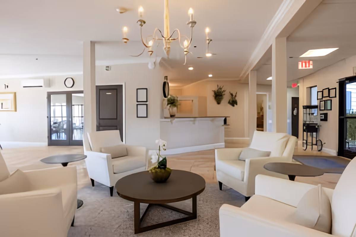 Bright senior living lounge with white armchairs arranged around round coffee tables and a reception desk in the background.