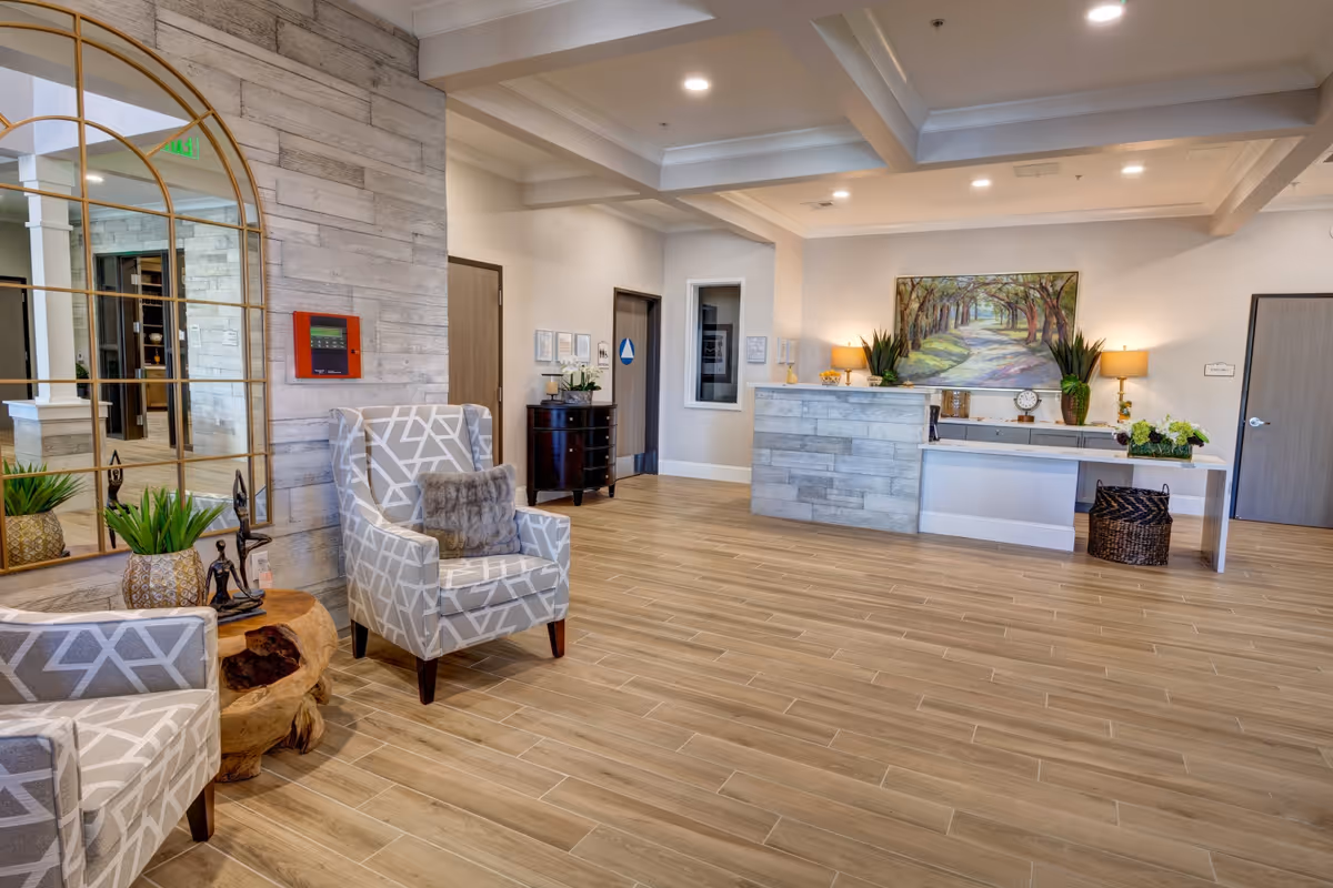 A bright and spacious reception area with light wood flooring and a modern stone reception desk. Two patterned armchairs with a small wooden side table and decorative plants are positioned near a large arched mirror on the left. The walls are light-colored with recessed lighting in the ceiling. A painting of a tree-lined path hangs behind the reception desk, which is decorated with plants, a clock, and lamps. Several doors and a small cabinet are visible in the background.