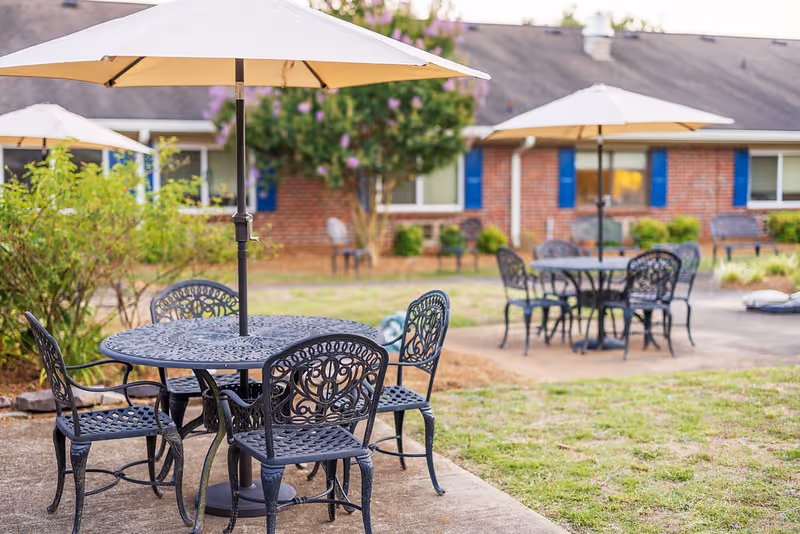Courtyard with wrought-iron patio tables and umbrellas on concrete paths in front of a brick residential building.