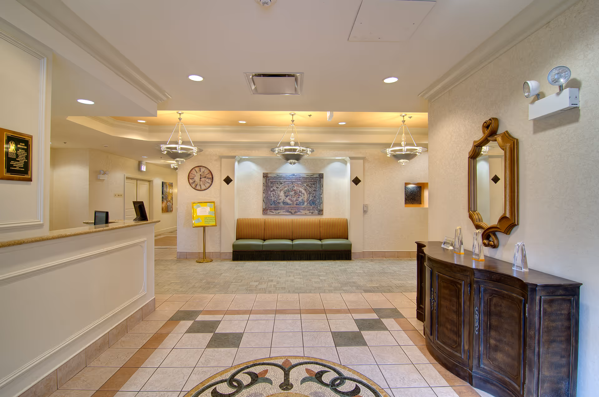 A well-lit reception area of Plum Creek Chicago featuring a front desk on the left, a decorative wooden console table with a mirror on the right, and a seating area with a striped bench and wall art in the background. The floor has a patterned tile design and there are three hanging light fixtures on the ceiling.