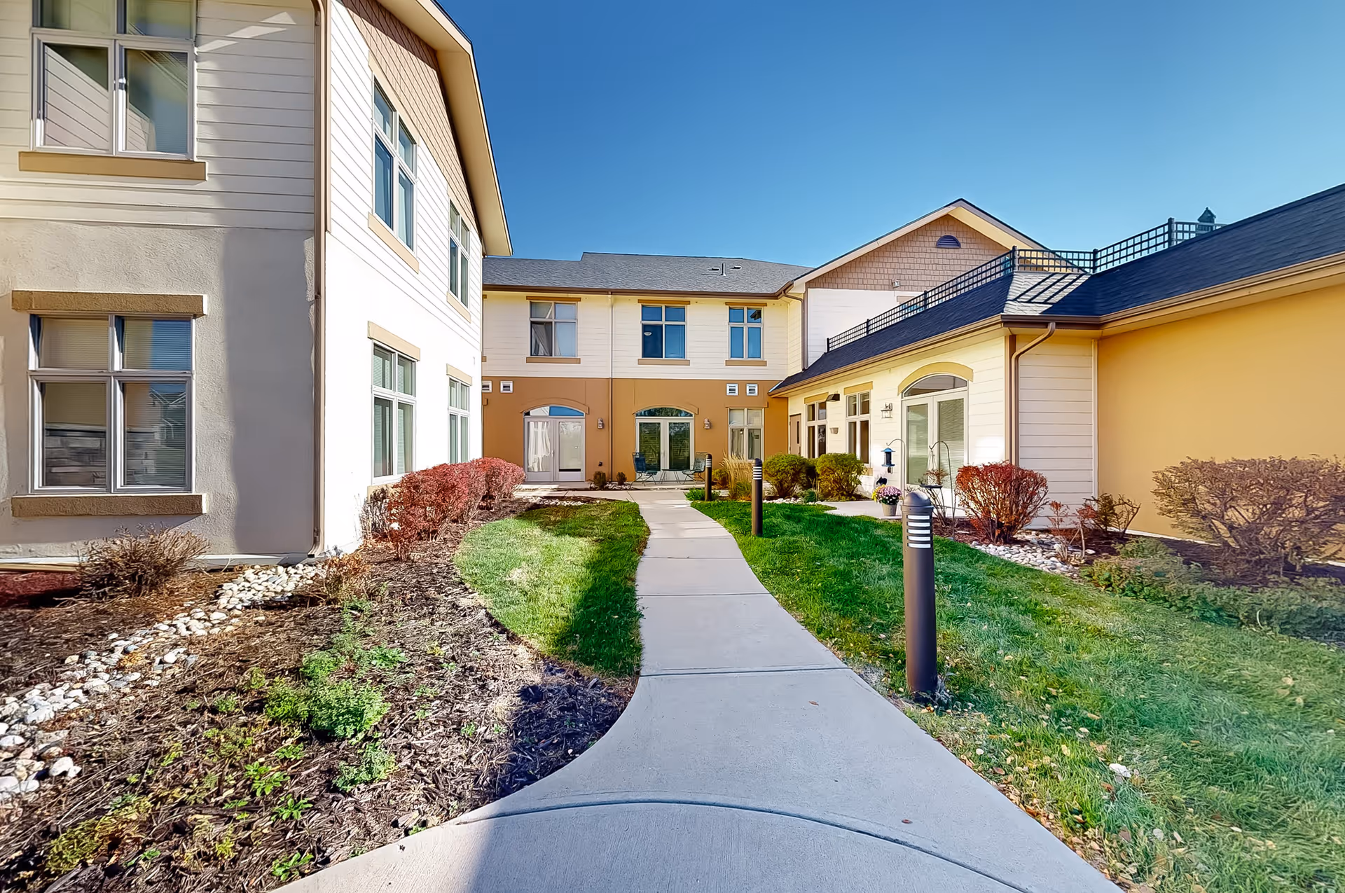 Outdoor courtyard area at MeadowView of Greeley featuring a curved concrete walkway surrounded by landscaped garden beds with shrubs and grass, bordered by a two-story building with multiple windows under a clear blue sky.