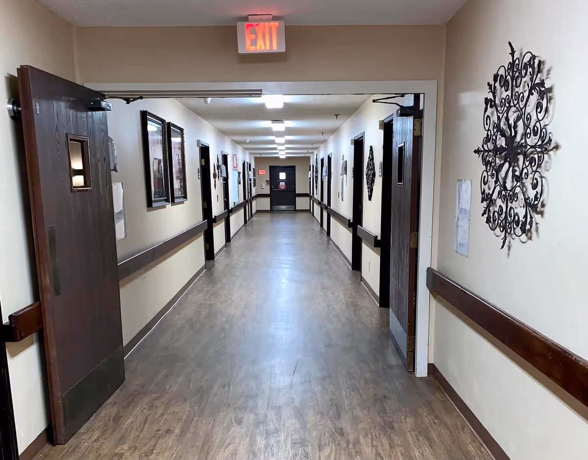 Long, well-lit nursing home corridor with wood doors, handrails, wall decorations and an EXIT sign.