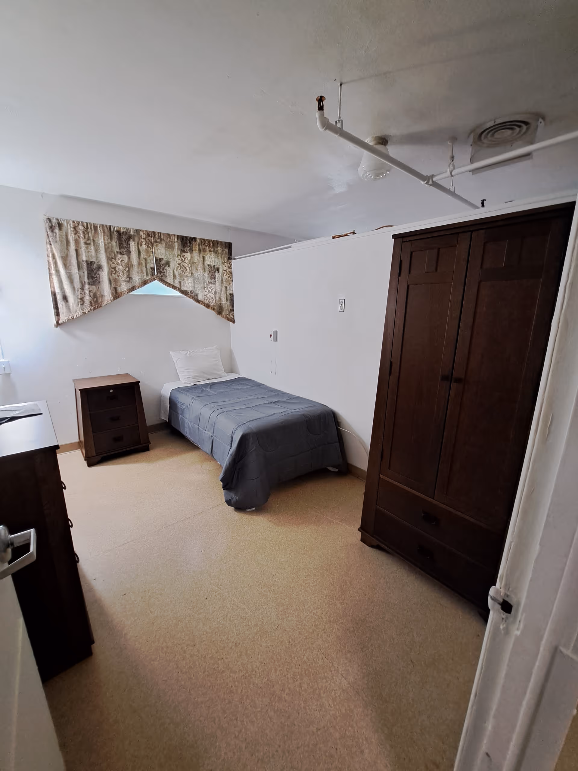 Small single-occupancy bedroom with a single bed, nightstand, and dark wooden wardrobe beneath a curtained window.