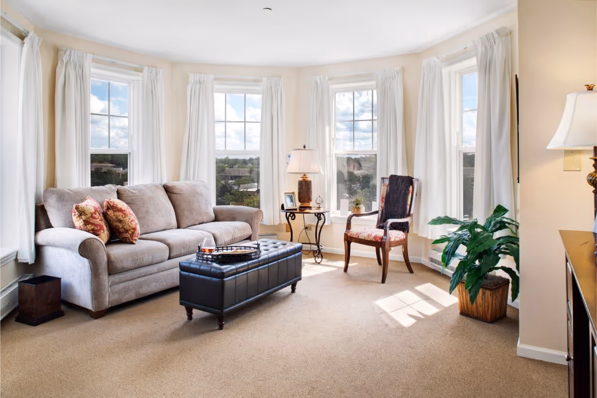 A bright living room with large windows covered by white curtains letting in natural light. The room features a beige sofa with decorative pillows, a black tufted ottoman with a tray on top, a wooden armchair with floral upholstery and a dark throw, a side table with a lamp and framed photos, a green potted plant, and beige carpeted flooring.