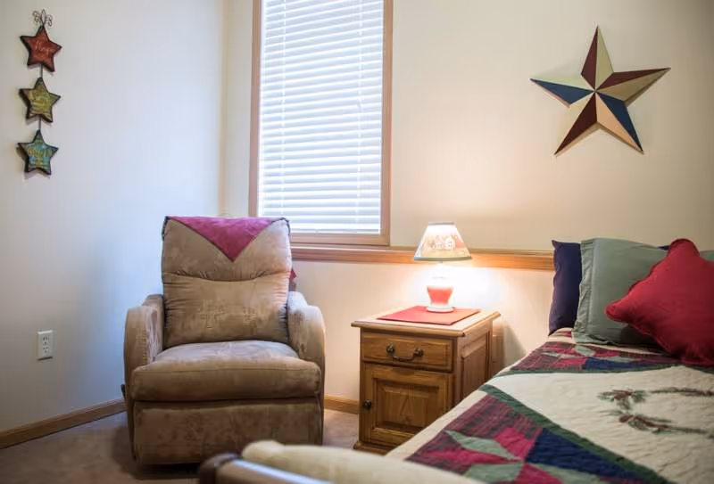 A cozy bedroom corner featuring a beige armchair with a purple throw draped over the back, a wooden nightstand with a small lamp on top, and part of a bed with a colorful quilt and multiple pillows. The walls are light-colored with a window covered by blinds and decorative star wall hangings.