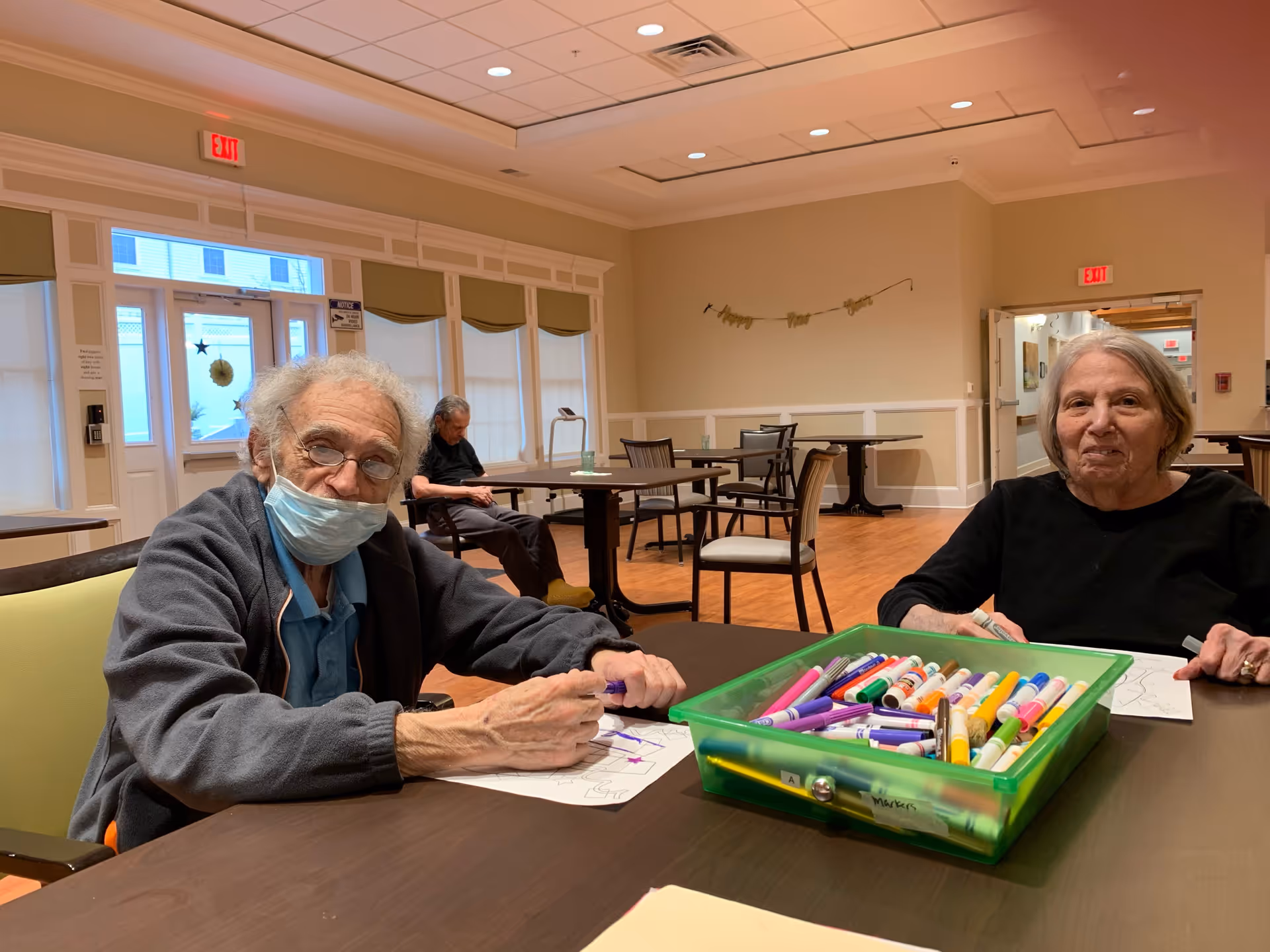 Two elderly individuals sitting at a table in a well-lit common area, engaging in coloring activities with a green container full of colorful markers in front of them. Another elderly person is seated in the background near windows with blinds. The room has wooden floors, beige walls, and multiple tables and chairs.