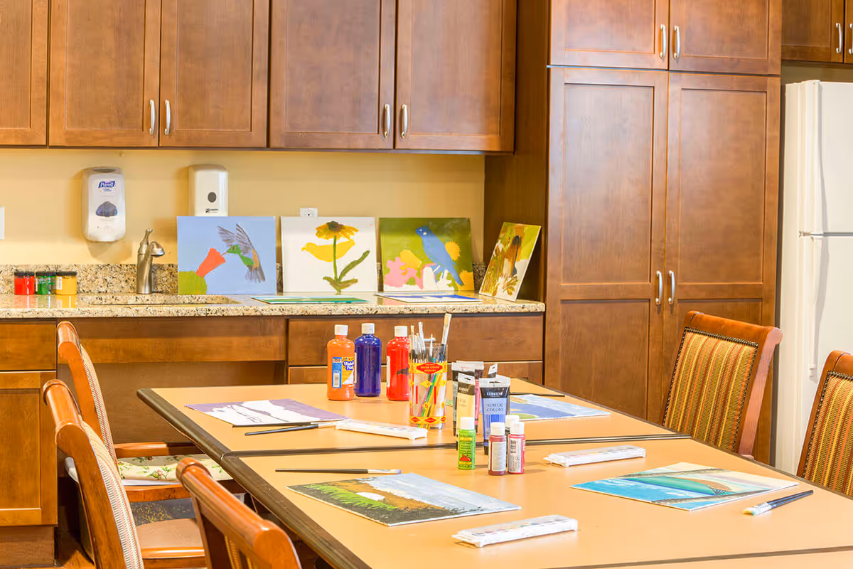 A craft room with a table set up for painting activities, featuring paint bottles, brushes, and canvases with colorful paintings of birds and flowers. The room has wooden cabinets and a granite countertop in the background.