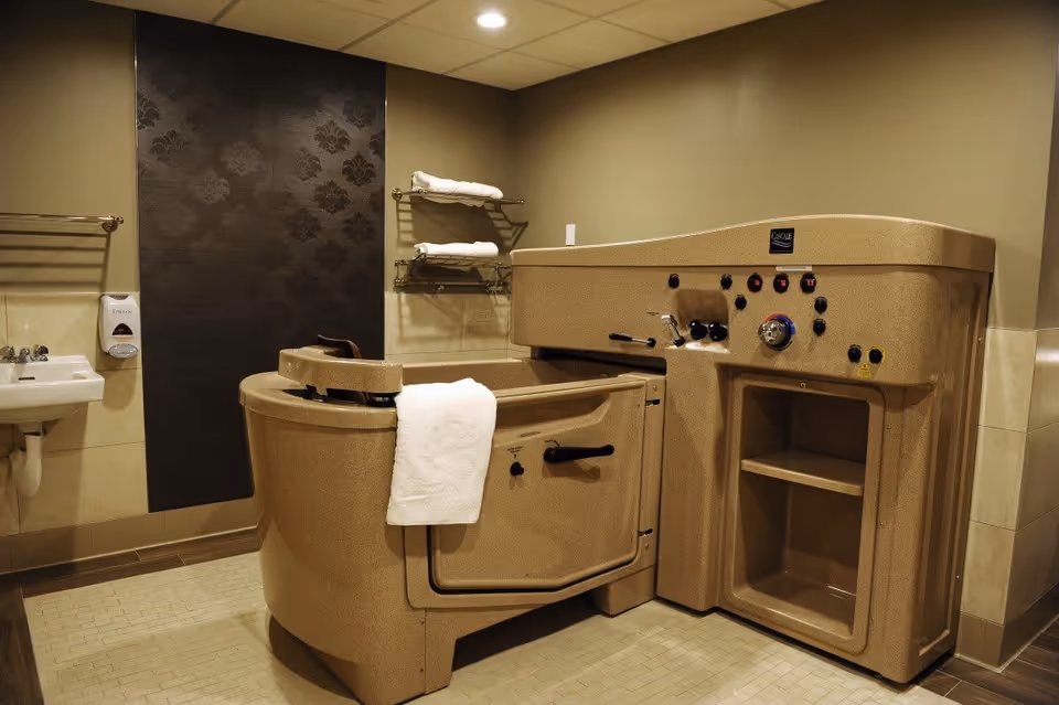 A beige-colored assisted bathing tub with control knobs and a door for easy access in a bathroom. The room has beige walls, a white sink with a soap dispenser, towel racks with folded white towels, and a dark decorative panel on one wall.