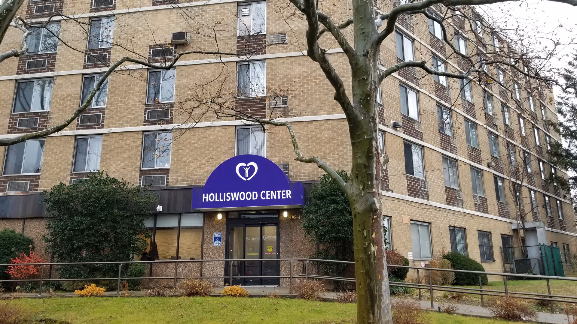 Multi-story brick building with a purple awning labeled "HOLLISWOOD CENTER" above the entrance and a tree in the foreground.
