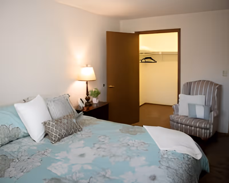A bedroom featuring a bed with patterned teal bedding, a nightstand and lamp, an open closet, and an upholstered armchair.