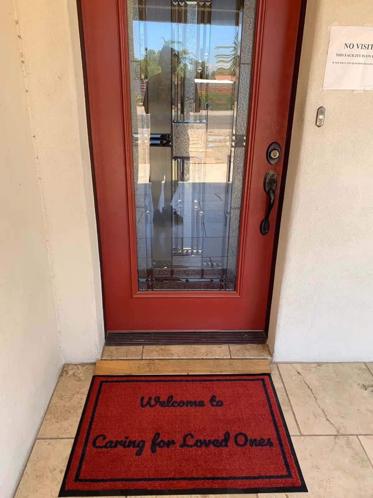 A red front door with decorative glass panels and a black handle. In front of the door is a red welcome mat with black text that reads 'Welcome to Caring for Loved Ones'. The door is set in a beige wall with tiled flooring. A small sign is partially visible on the right side of the door.