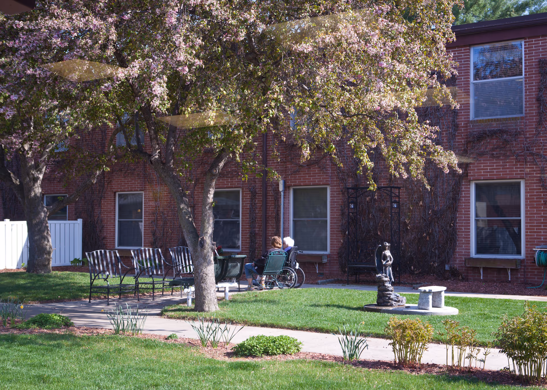 Courtyard outside a brick nursing facility with flowering trees, lawn, benches, a small fountain statue, and a person sitting in a wheelchair.