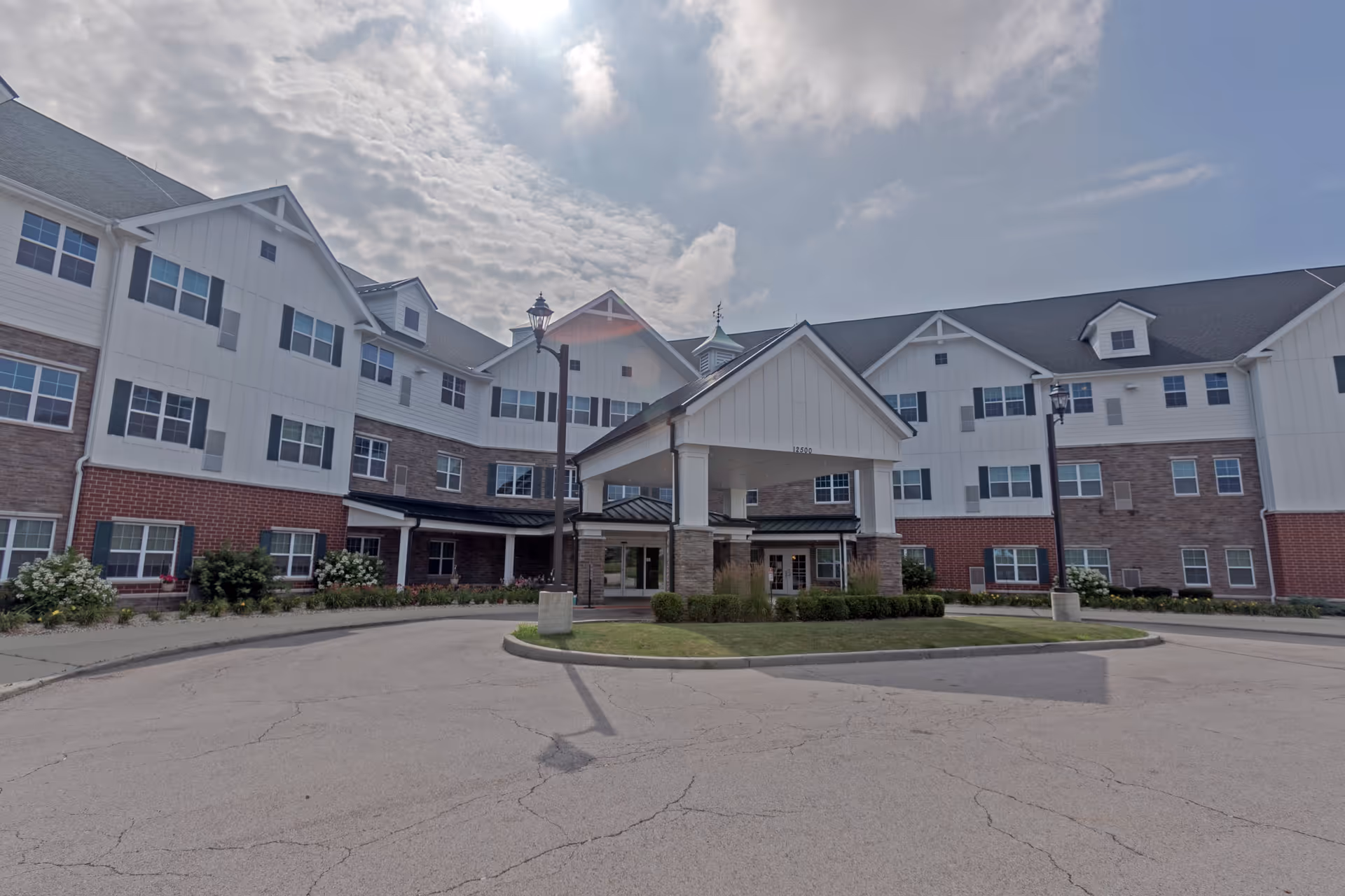 Front exterior view of a large senior living facility building with white siding, brick accents, multiple windows, and a covered entrance with columns. The sky is partly cloudy.