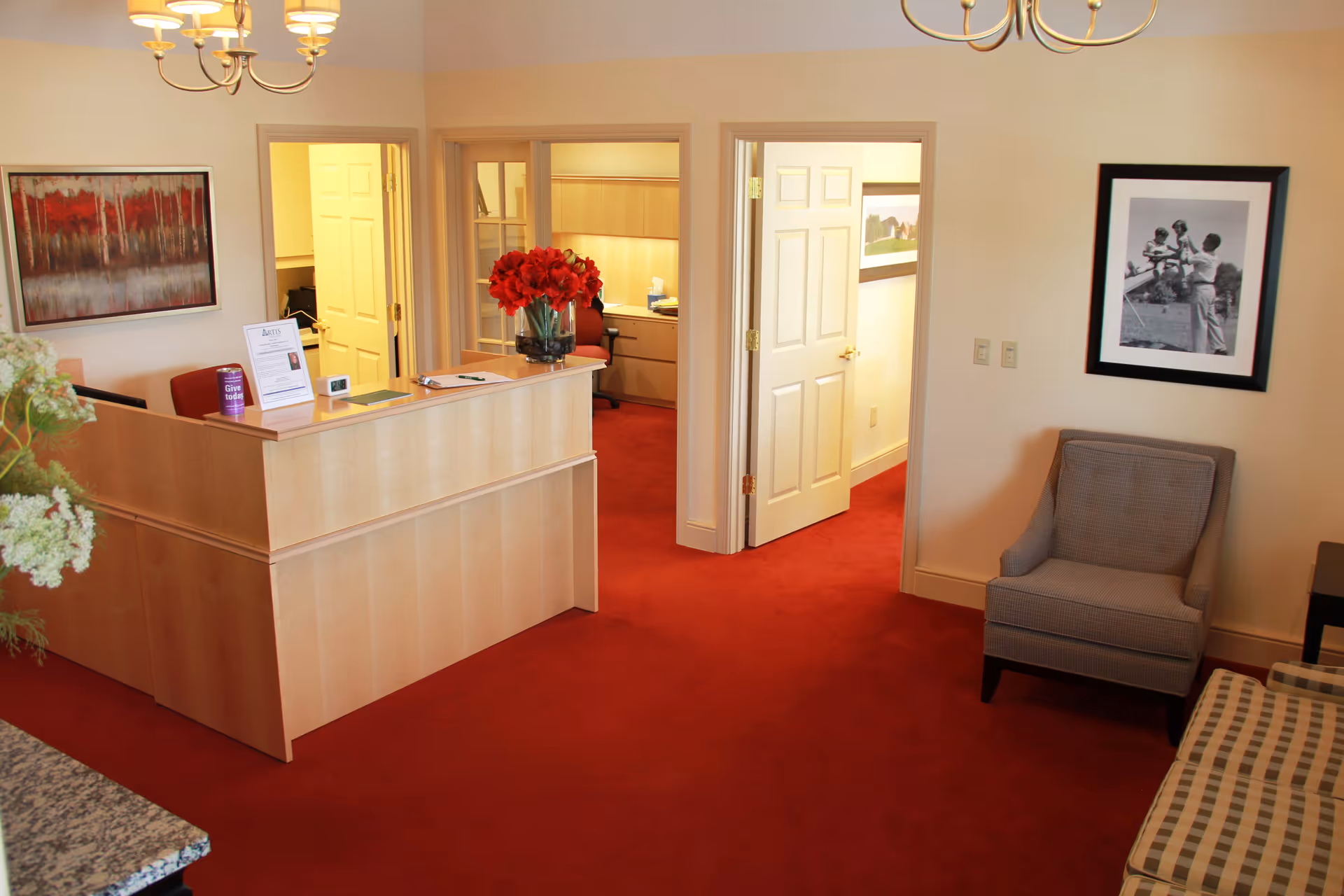 Reception area with a wooden desk adorned with a vase of red flowers, a red carpeted floor, a gray armchair, a checkered sofa, and framed artwork on the walls. Two open doors lead to adjoining rooms with office furniture visible.
