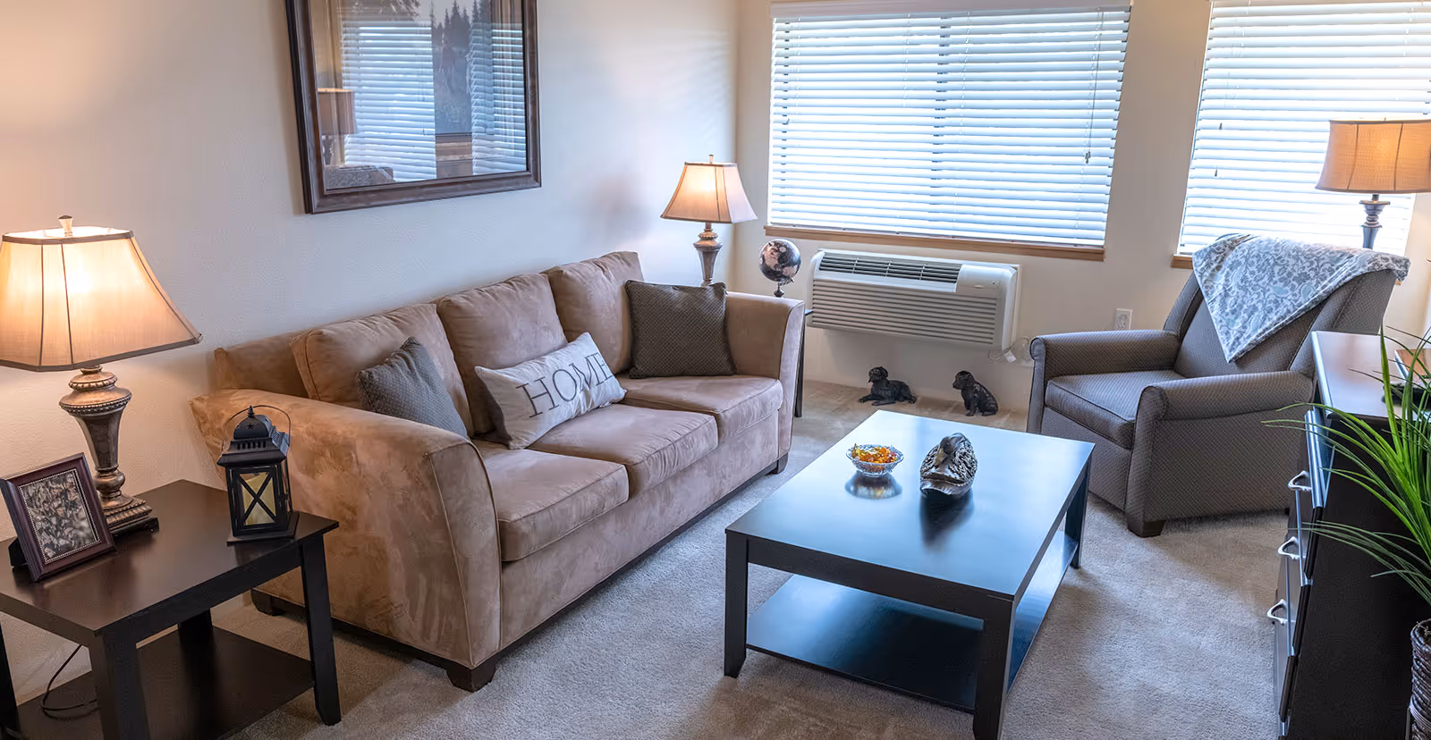 Bright living room with a beige sofa, armchair, coffee table, lamps, and window blinds.