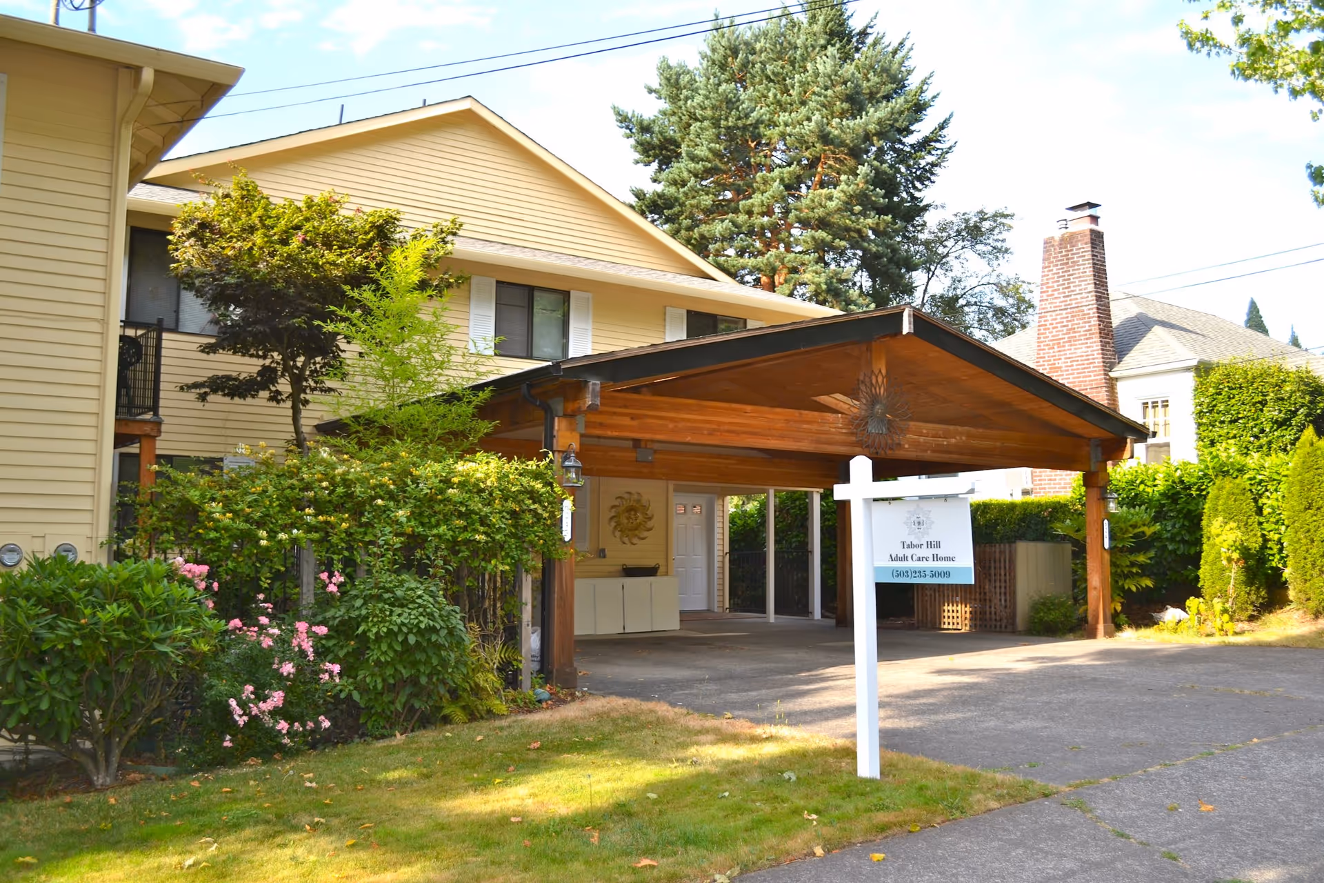 Exterior view of Tabor Hill Care Homes showing a two-story beige building with white shutters, a wooden carport, green bushes, trees, and a lawn in front. A white signpost with the facility name and phone number is visible near the driveway.