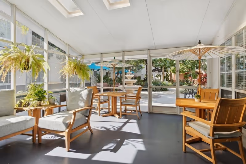 A bright and airy sunroom with wooden chairs and tables, some with cushions, and hanging green plants. The room has large windows and glass doors that open to an outdoor garden area with a fountain and greenery.