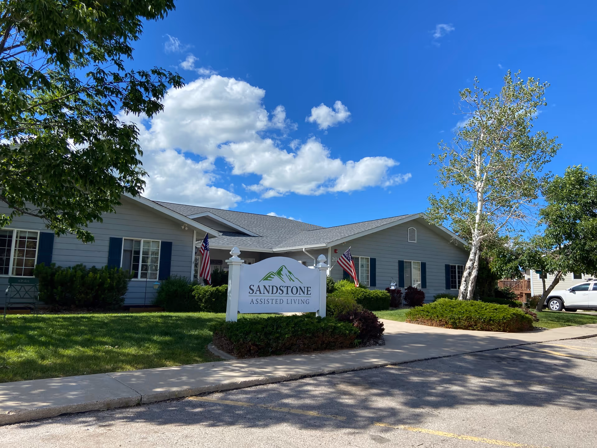 Exterior view of Sandstone Assisted Living facility on a sunny day with blue sky and scattered clouds. The building is a single-story structure with gray siding, white trim, and blue shutters. There are two American flags displayed near the entrance. A white sign with green mountain graphics and the text 'Sandstone Assisted Living' is prominently placed in front of the building, surrounded by green bushes and trees.