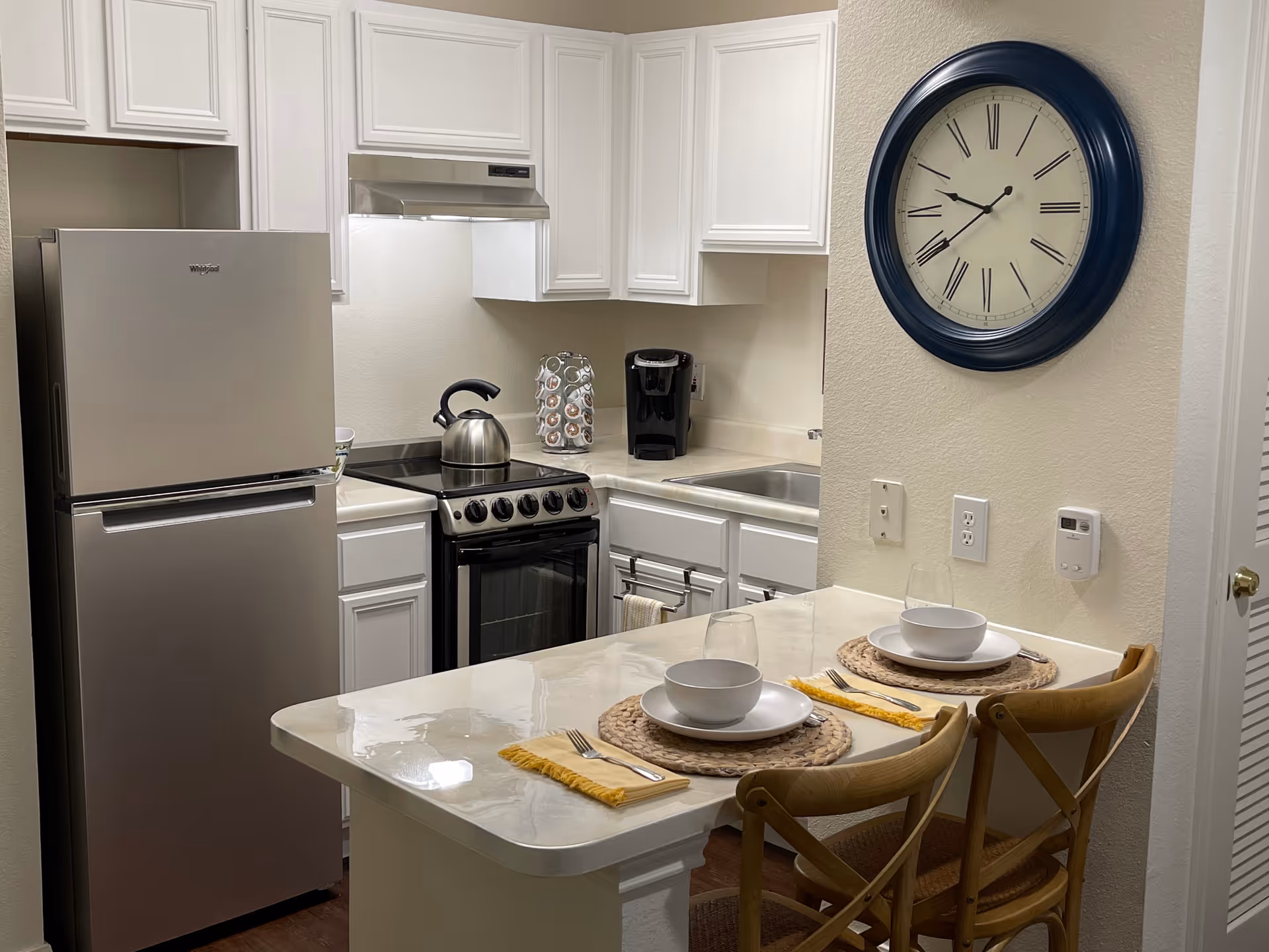 Small kitchen with stainless steel refrigerator and stove, white cabinets, a breakfast bar set for two, and a large wall clock.