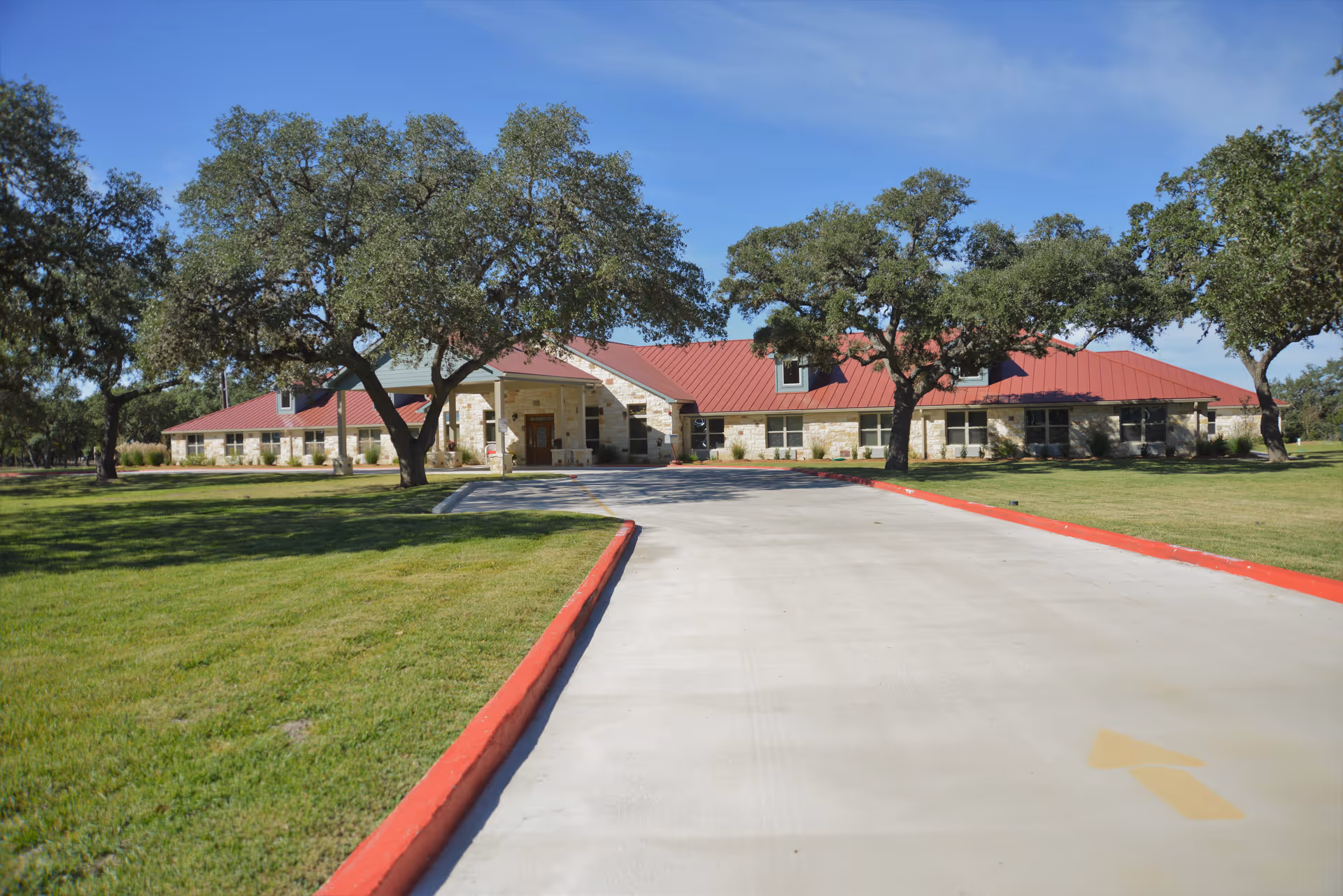 Front exterior view of Gemstone Senior Living at Bulverde, a single-story building with a red roof, stone facade, surrounded by green grass and trees under a clear blue sky.