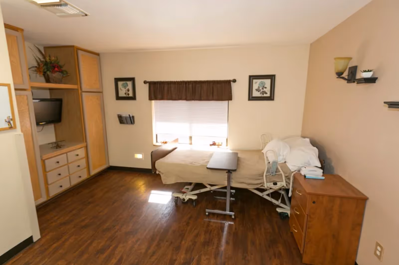 Private senior bedroom with an adjustable hospital bed, bedside table, built-in cabinets and a wall-mounted TV.